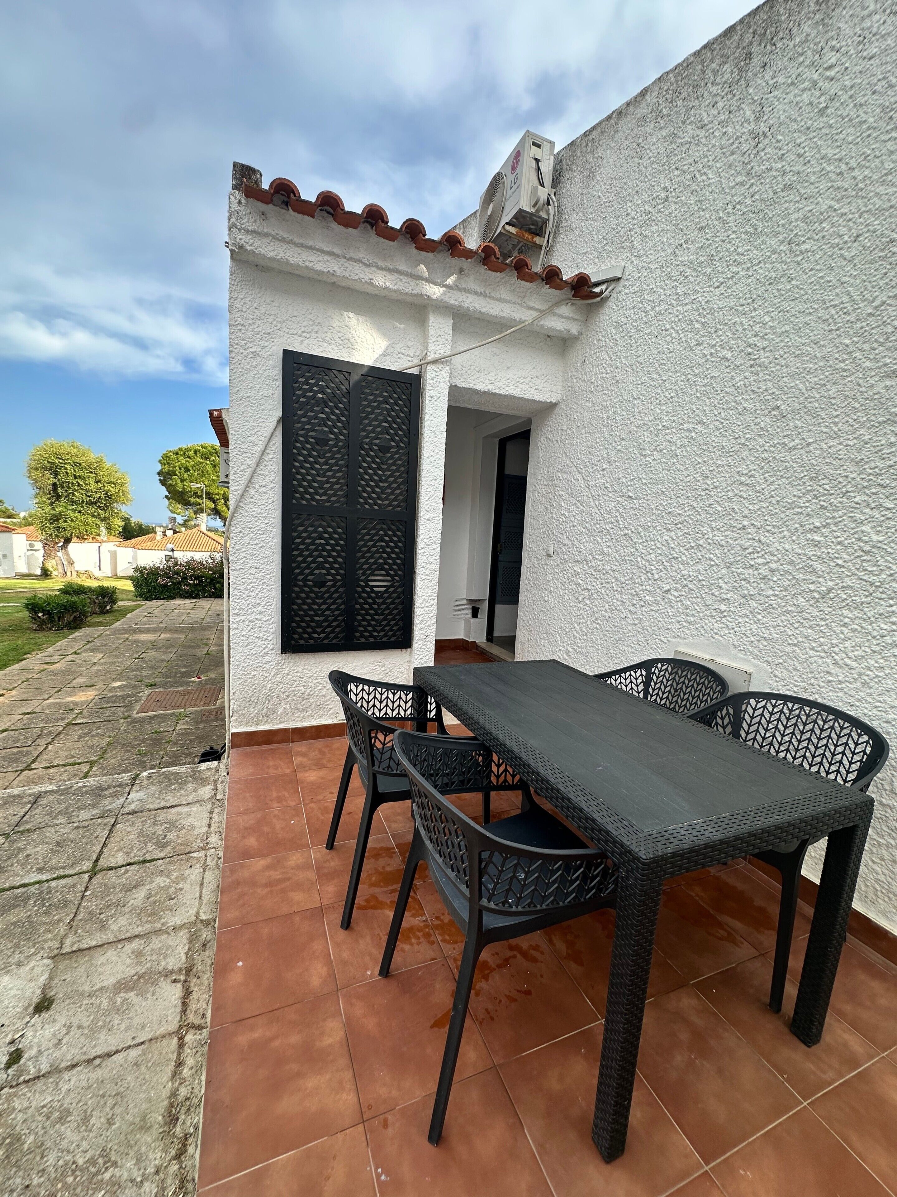 Uma área de pátio com mesa e cadeiras, uma parede branca e piso ladrilhado.