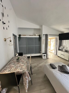 A compact room with a dining table, chairs, a bed, a TV, and a window with curtains.