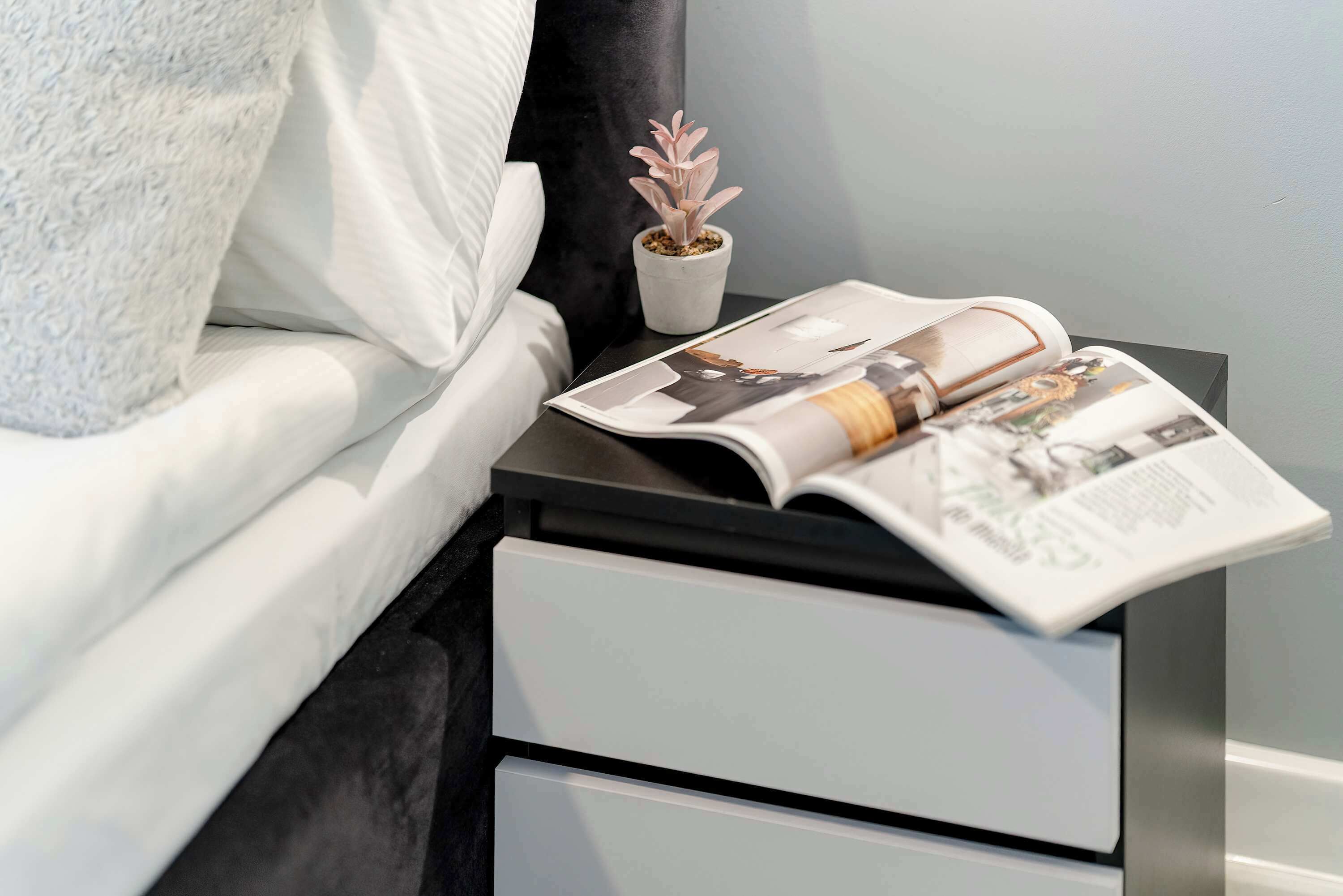A neatly made bed with a pillow, a small potted plant, and a magazine on a bedside table.