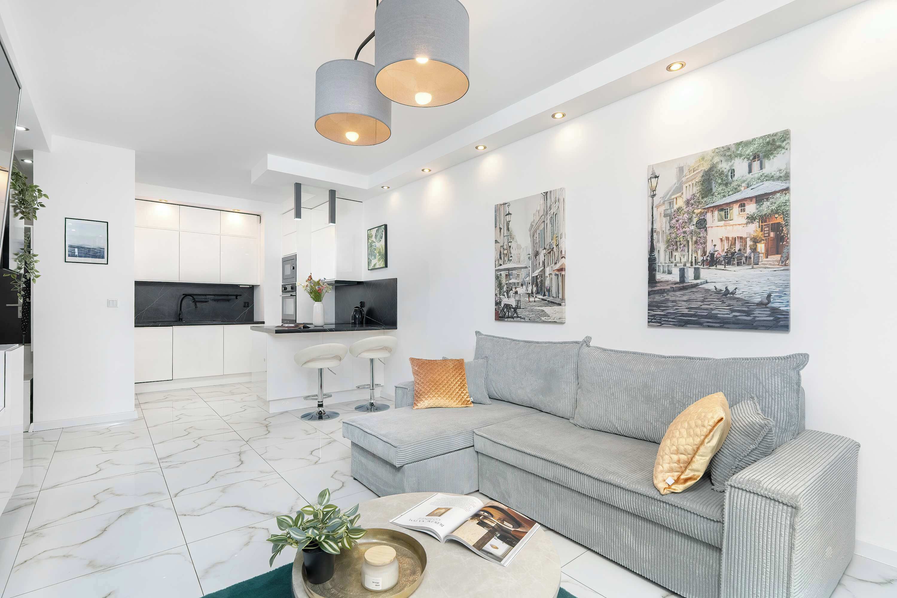 Un salon moderne avec un canapé gris, une table basse sur laquelle repose un livre, et une cuisine en arrière-plan.