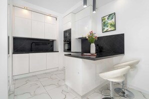 A modern kitchen with white cabinetry, a black countertop, a stainless steel sink, a microwave, and a bar stool. - Modern Apartment with a Balcony | Quiet Neighborhood in Poznan (Poznań)