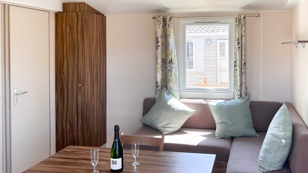 Een kamer met een houten kledingkast, een bank met kussens, een tafel met een fles en glazen, en een raam met gordijnen.