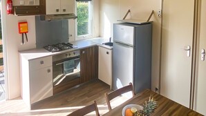 Een functionele keuken met een fornuis, oven en koelkast, een eettafel met stoelen, en een raam voor natuurlijk licht.