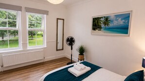 Et soveværelse med en seng, et vindue, en radiator, en ventilator, en plante og et billede af en strand.