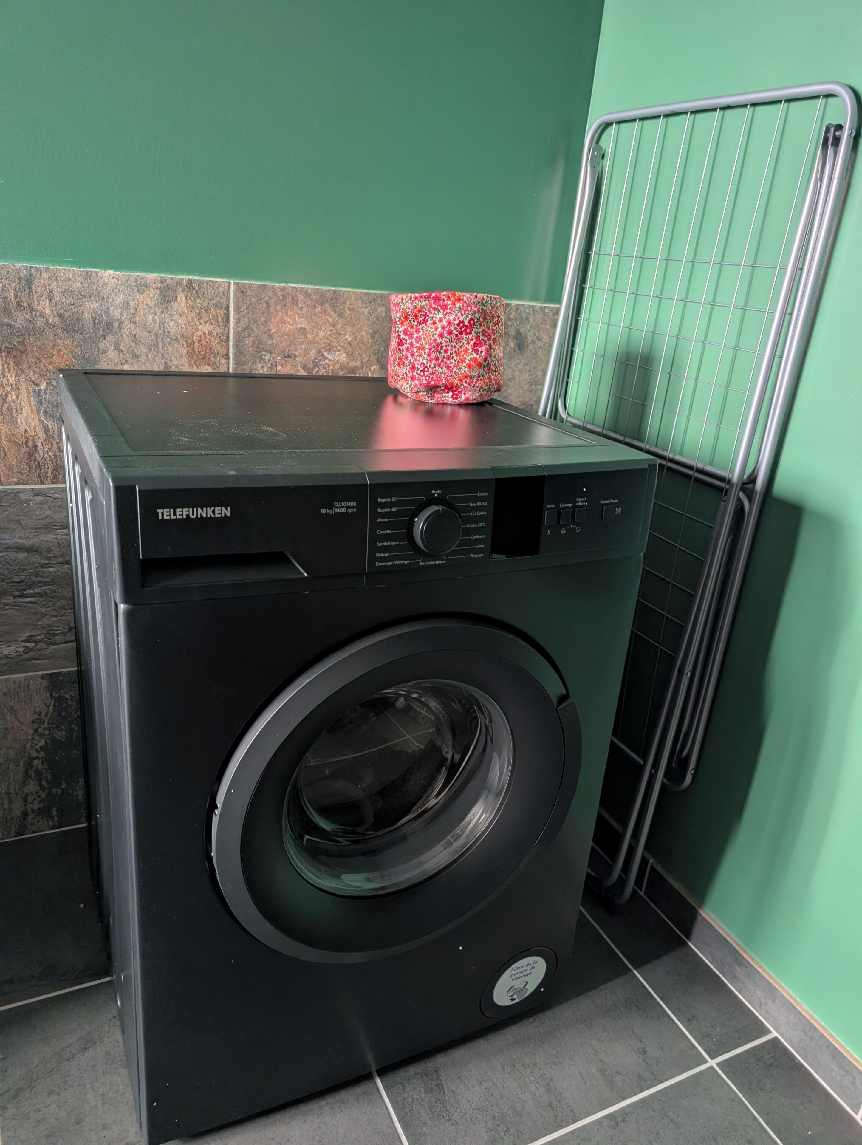 A front-loading washing machine with a control panel, placed in a room with a tiled floor and a green wall.