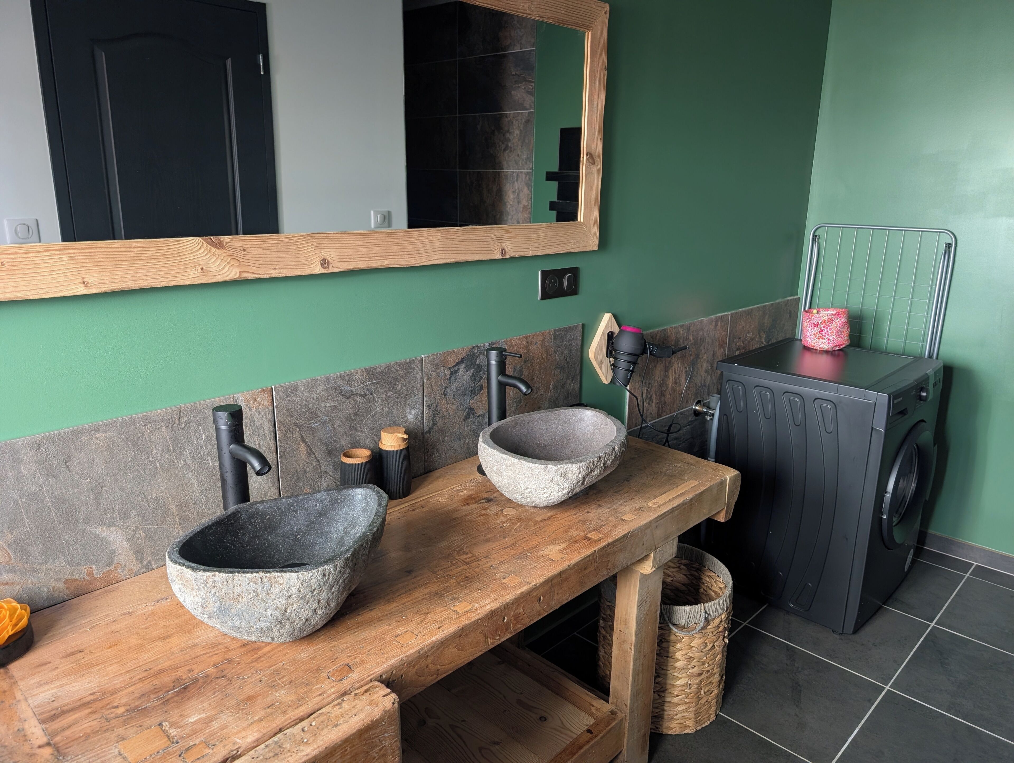Ein Badezimmer mit einem Holzbadezimmerschrank, zwei Steinwaschbecken, einem Spiegel, einer schwarzen Armatur und einer Waschmaschine, sowie einer grünen Wand.