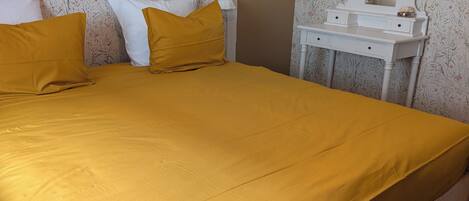 A bed with a mustard-colored bedspread, a white headboard, and a bedside table with a mirror and a lamp.