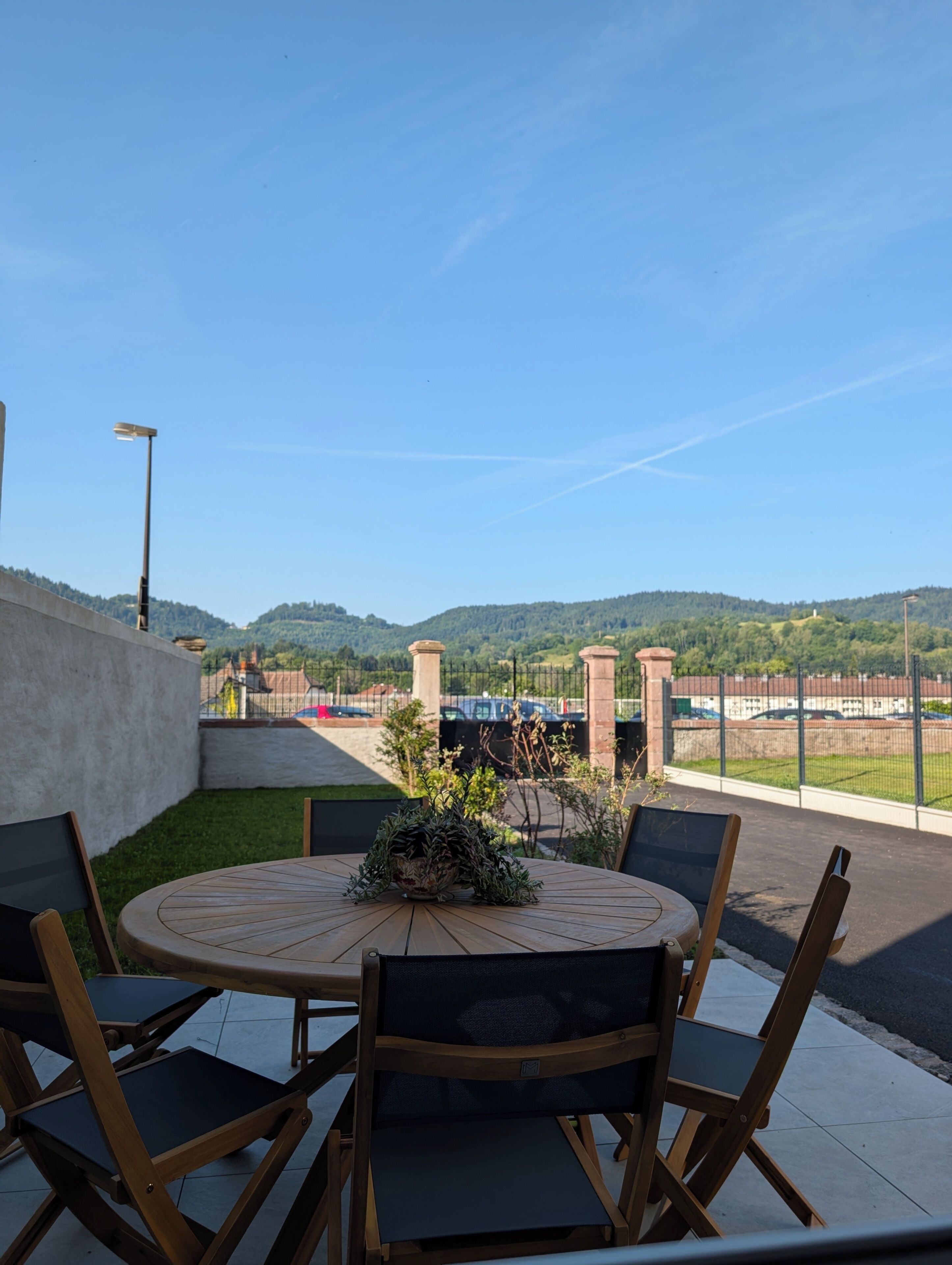 Une terrasse extérieure avec une table ronde et des chaises, un pot de fleurs et une vue sur un parking et des collines en arrière-plan.