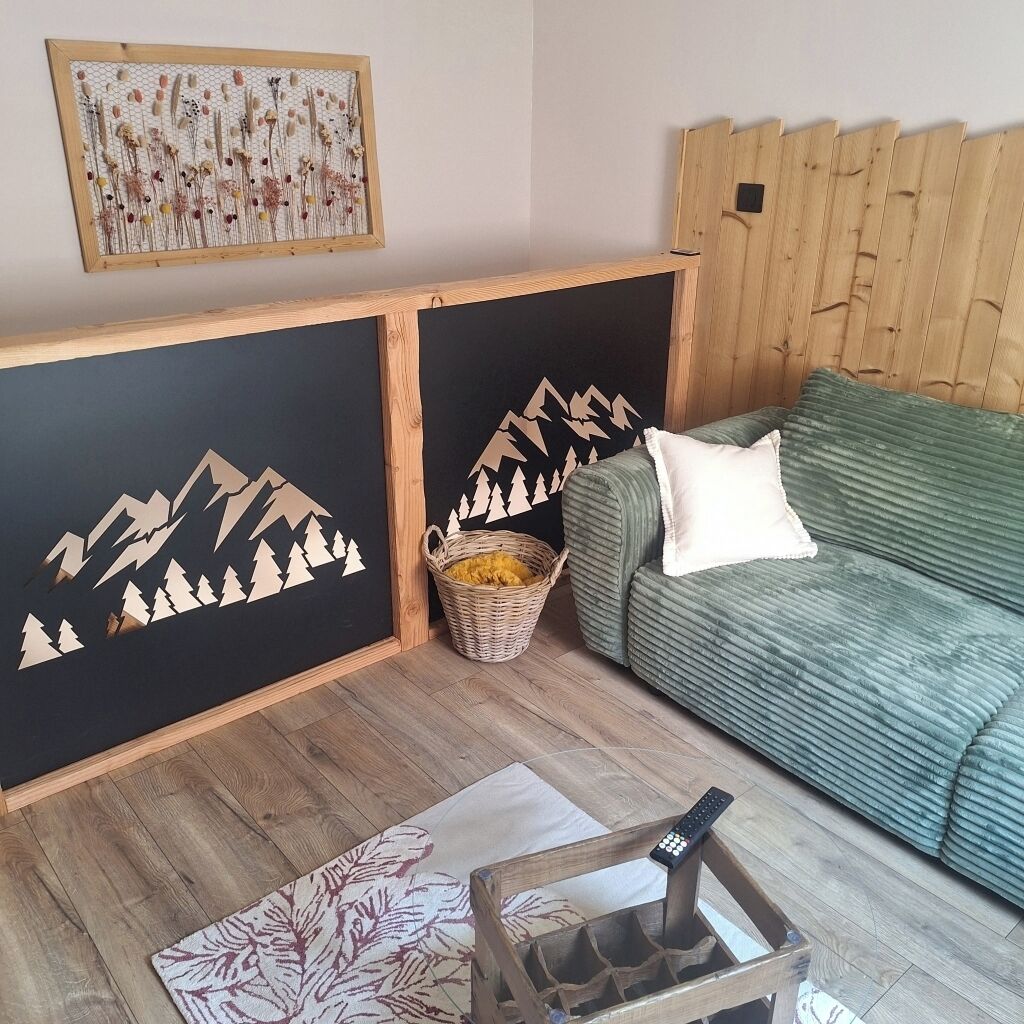 A room with a wooden headboard, a green sofa, a wall with mountain and forest decals, a wicker basket, and a remote control on a wooden stand.