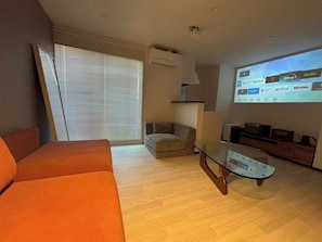 Ett modernt vardagsrum med en soffa, ett soffbord i glas, en TV som är väggmonterad ovanför en platt-TV och ett fönster med persienner.