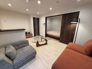 Een knus woongedeelte met een bank, een glazen salontafel, een bed en een bureau met een lamp.