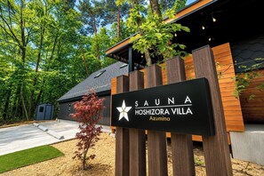 Schild für 'SAUNA HOSHIZORA VILLA Azumino' mit einem Stern-Logo, umgeben von Bäumen und einem Gebäude mit Holzfassade.