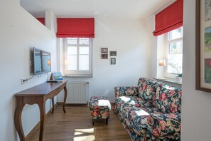 Ein Zimmer mit einem Holztisch, einem blumigen Sofa, einem Fernseher und einem Fenster mit roten Vorhängen.
