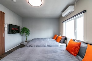 Ein modernes Schlafzimmer mit einem Bett, einer Topfpflanze, einem an der Wand montierten Fernseher und einem Fenster mit Jalousien.
