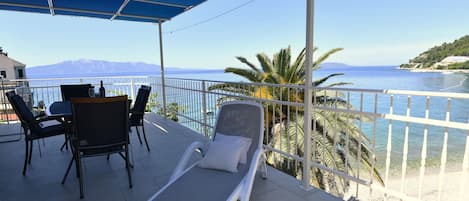 A balcony with a table and chairs, a sun lounger, and a view of the sea and mountains.