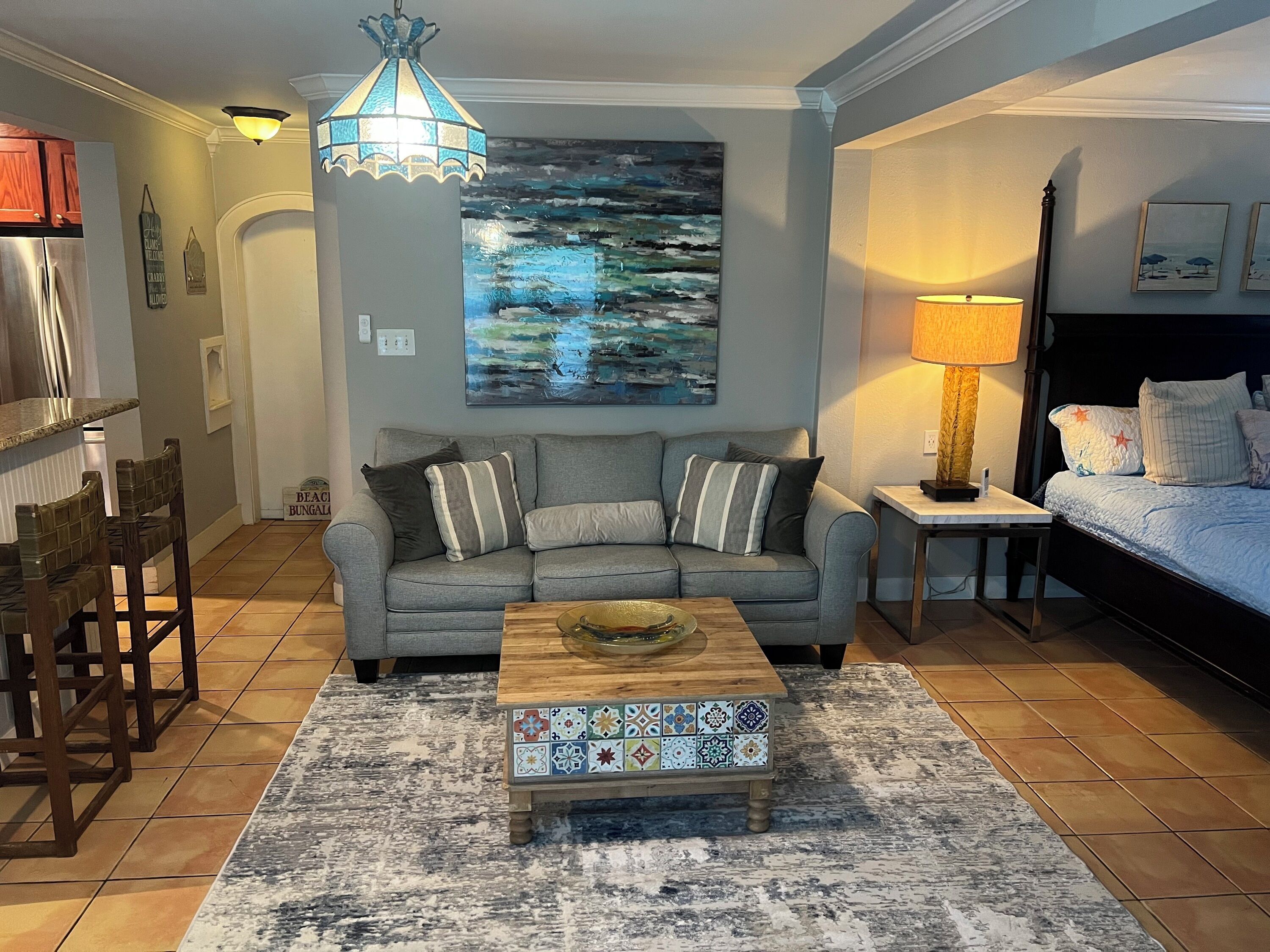 A living room with a grey sofa, a coffee table, a painting, a lamp, and a bed with a headboard.