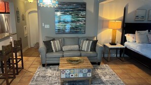 A living room with a grey sofa, a coffee table, a painting, a lamp, and a bed with a headboard. - Paradise Pool House (Galveston)