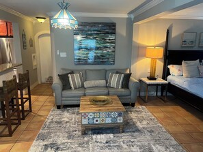 A living room with a grey sofa, a coffee table, a painting, a lamp, and a bed with a headboard. - Paradise Pool House (Galveston)