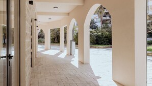 Uma galeria coberta com arcos, piso de azulejos e vista para uma área residencial.
