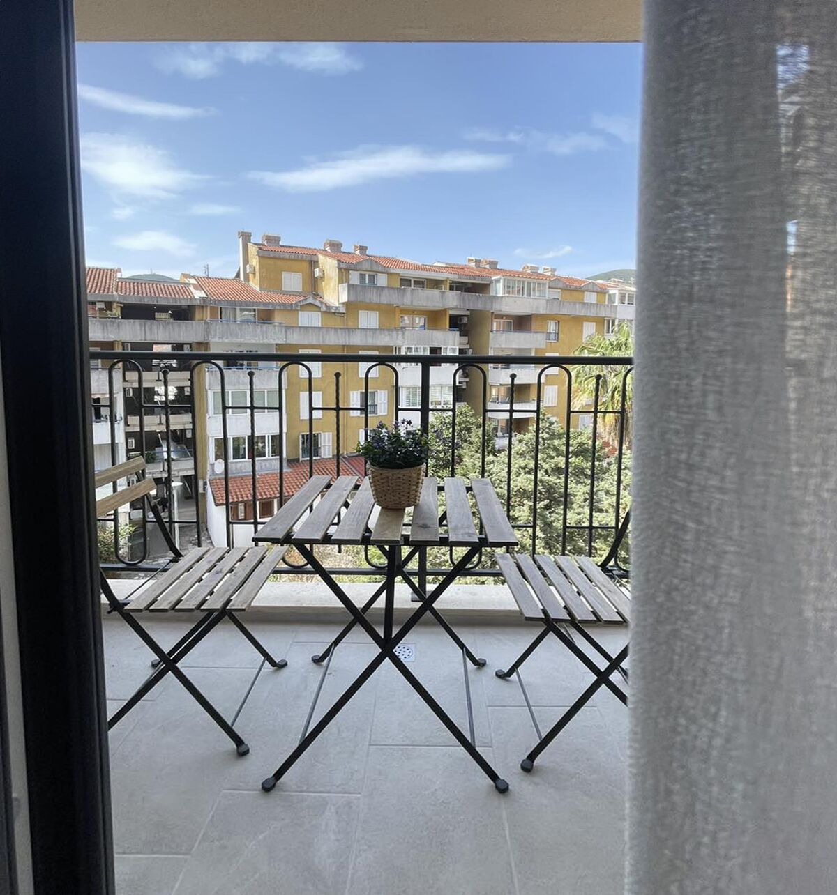 A balcony with a wooden table and chairs, a potted plant, and a view of residential buildings.
