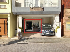 Un edificio con zona de estacionamiento techada, un coche aparcado en su interior y una entrada visible con puertas de cristal.