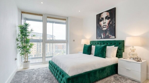 Une chambre à coucher avec un lit recouvert d’un tissu vert, une grande fenêtre, une plante en pot et un tableau accroché au mur.