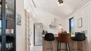 A modern kitchen with a stainless steel refrigerator, wooden bar stools, and a window providing natural light. - 5 Star Holiday Home in Nexo (Nexø)