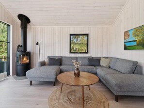 Ein modernes Wohnzimmer mit einer Eckcouch, einem runden Holztisch, einem Kamin und einem großen Fenster mit Blick auf Bäume.