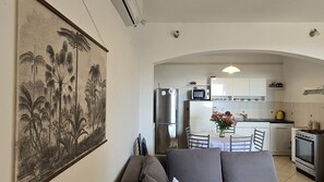 Eine moderne Küche mit Kühlschrank, Herd und Mikrowelle. Ein Esstisch mit Stühlen und eine Blumenvase stehen bereit. Ein großes gerahmtes Bild ist an der Wand aufgehängt.