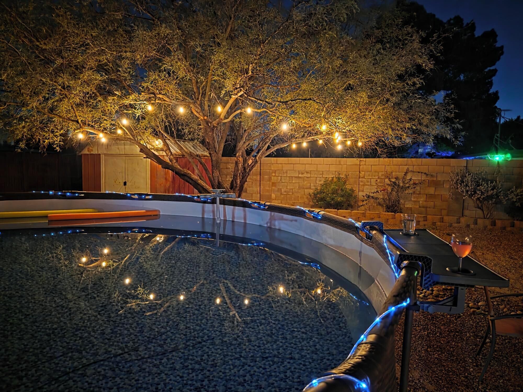 A swimming pool with a curved edge, surrounded by a brick wall and a tree with string lights.
