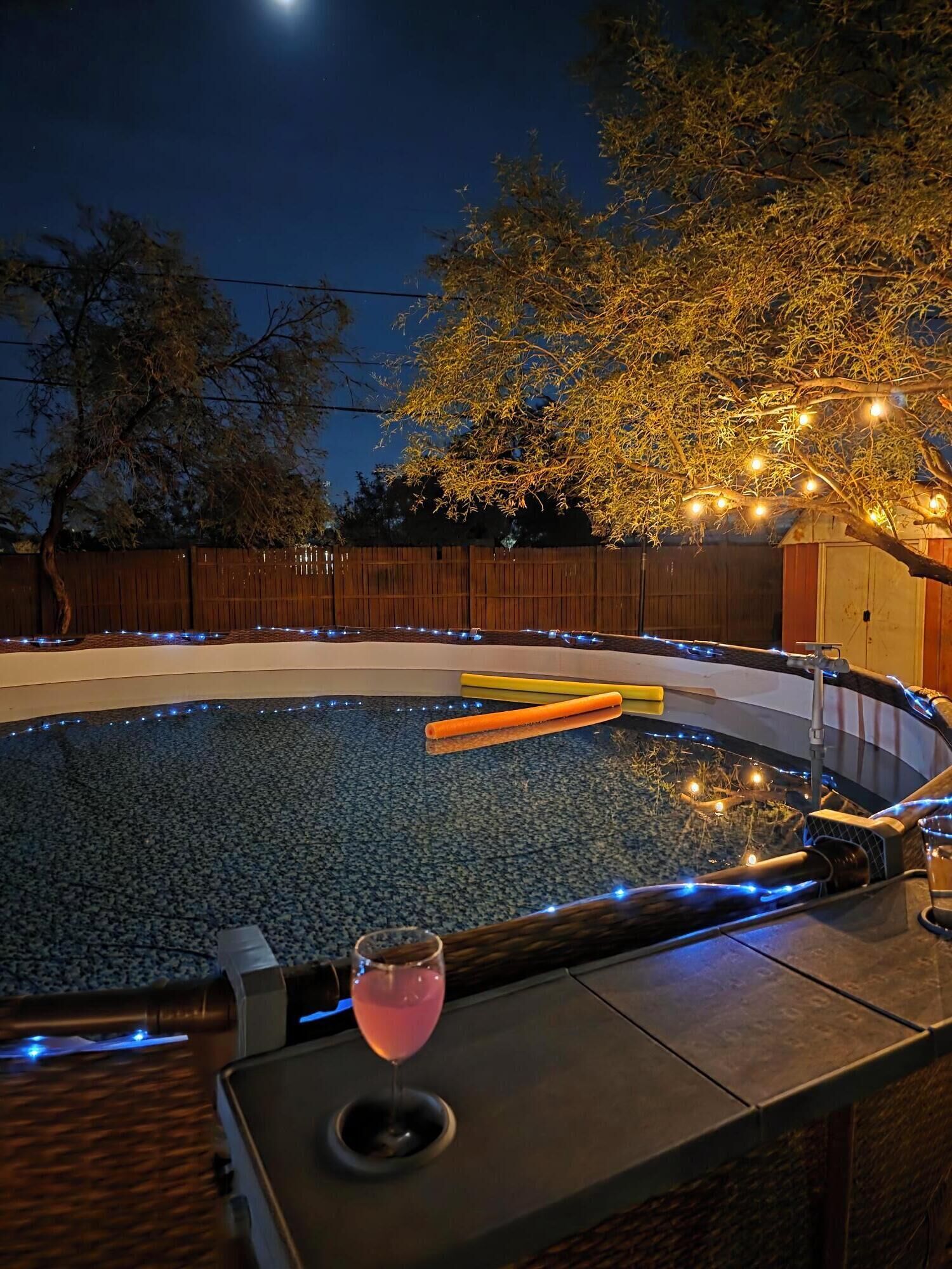 A hot tub with a glass of pink drink, surrounded by a wooden fence and string lights.