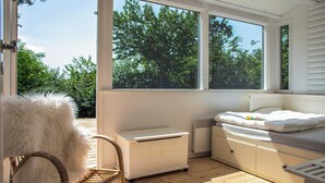 A room with a bed, a chair, and a small table, with large windows offering a view of trees. - Sea-view terrace on scenic Helgenæs (Knebel)