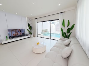A modern living room with a TV, a round coffee table, a white sofa, a large window with a view of a swimming pool, and a potted plant. - POOL Villa KTV BBQ Friends Thepprasit (Muang Pattaya)