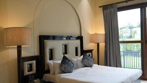 A hotel room with a large bed, two bedside tables, a bench, a window with curtains, and a view of the outdoors. - Rosewood Manor BNB (Alwar)