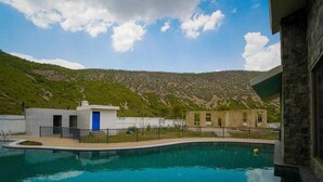 A swimming pool with a view of a mountainous landscape, a small building with a blue door, and a stone wall. - Rosewood Manor BNB (Alwar)