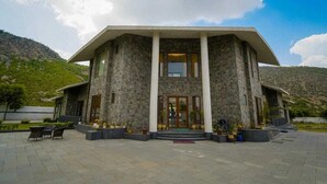 A large building with a stone facade, a central entrance with glass doors, and a paved courtyard with outdoor furniture. - Rosewood Manor BNB (Alwar)