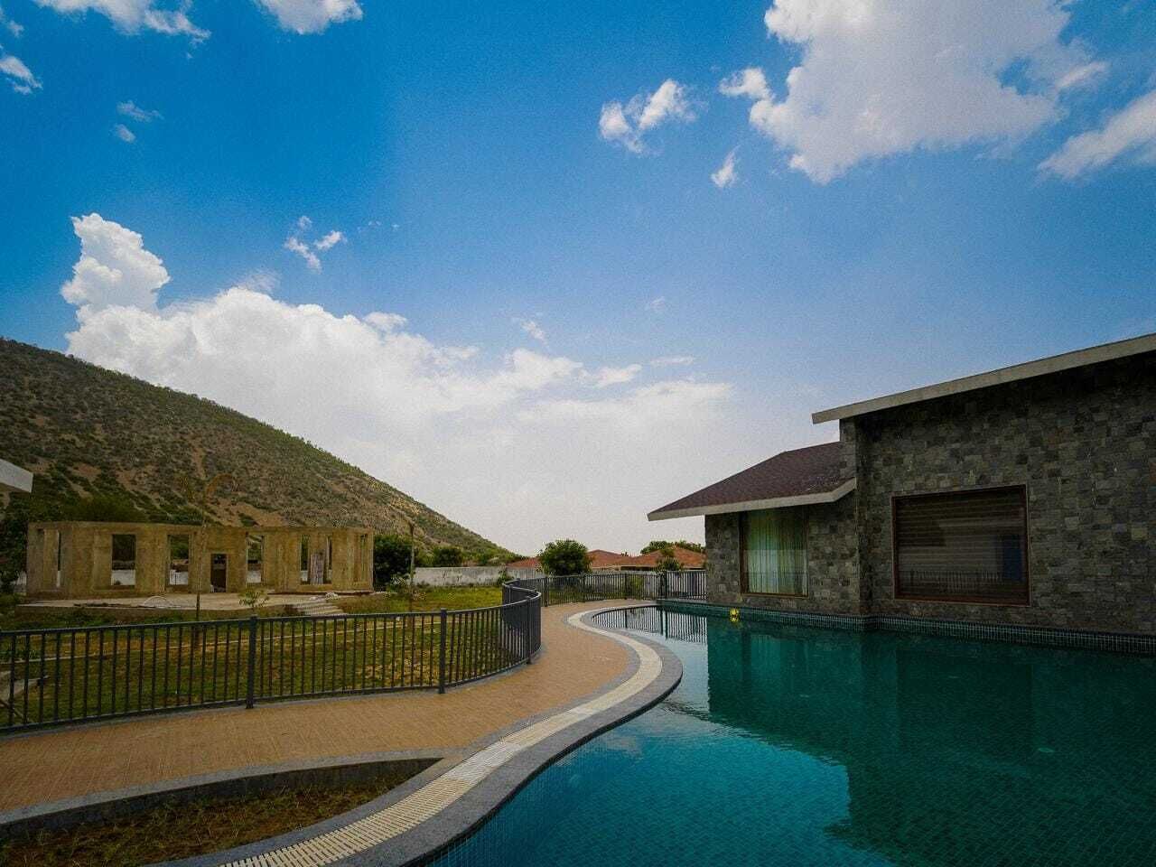 Zona de piscina amb un edifici de pedra, un passeig pavimentat i un paisatge amb turons al fons.