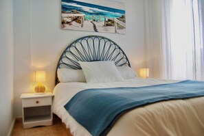 Ein ordentlich gemachtes Bett mit einer blauen Decke, einem weißen Kissen und einem Kopfteil mit einem kreisförmigen Design. Ein Nachttisch mit einer Lampe und ein Bild von einer Strandansicht an der Wand sind ebenfalls vorhanden.