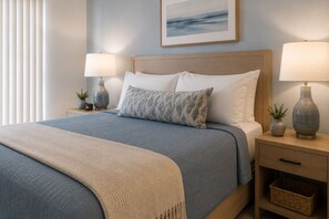 Una cama bien hecha con una colcha azul y beige, dos mesitas de noche con lámparas, un cuadro en la pared y una planta pequeña en la mesita.
