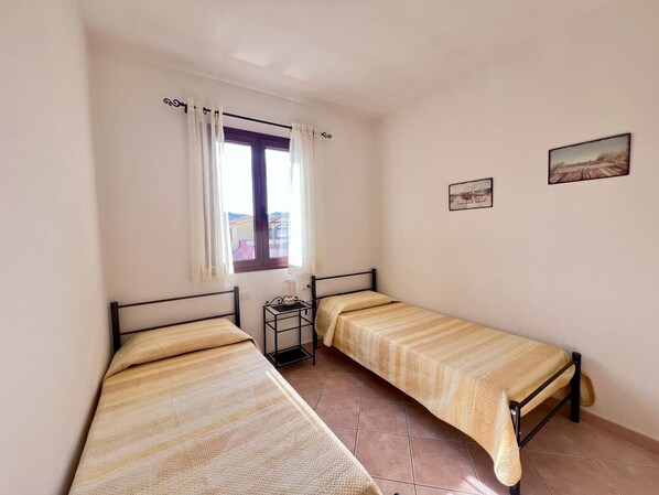Una habitación pequeña con dos camas individuales, una ventana con cortinas, una mesita con lámpara y dos cuadros en la pared.
