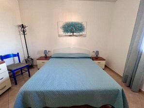 Een slaapkamer met een bed, een stoel, een nachtkastje, een lamp, een schilderij van een boom en een vloerlamp.