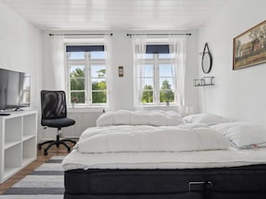 Ett sovrum med en säng, ett skrivbord med en tv, två fönster med gardiner och en tavla på väggen.