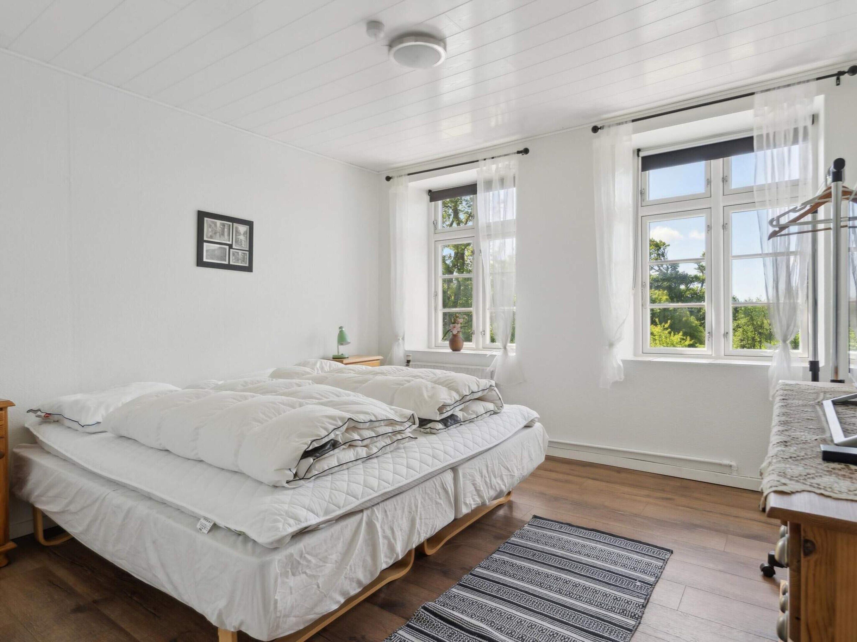 A bedroom with two beds, a bedside table, a lamp, a window with curtains, and a view of greenery outside.