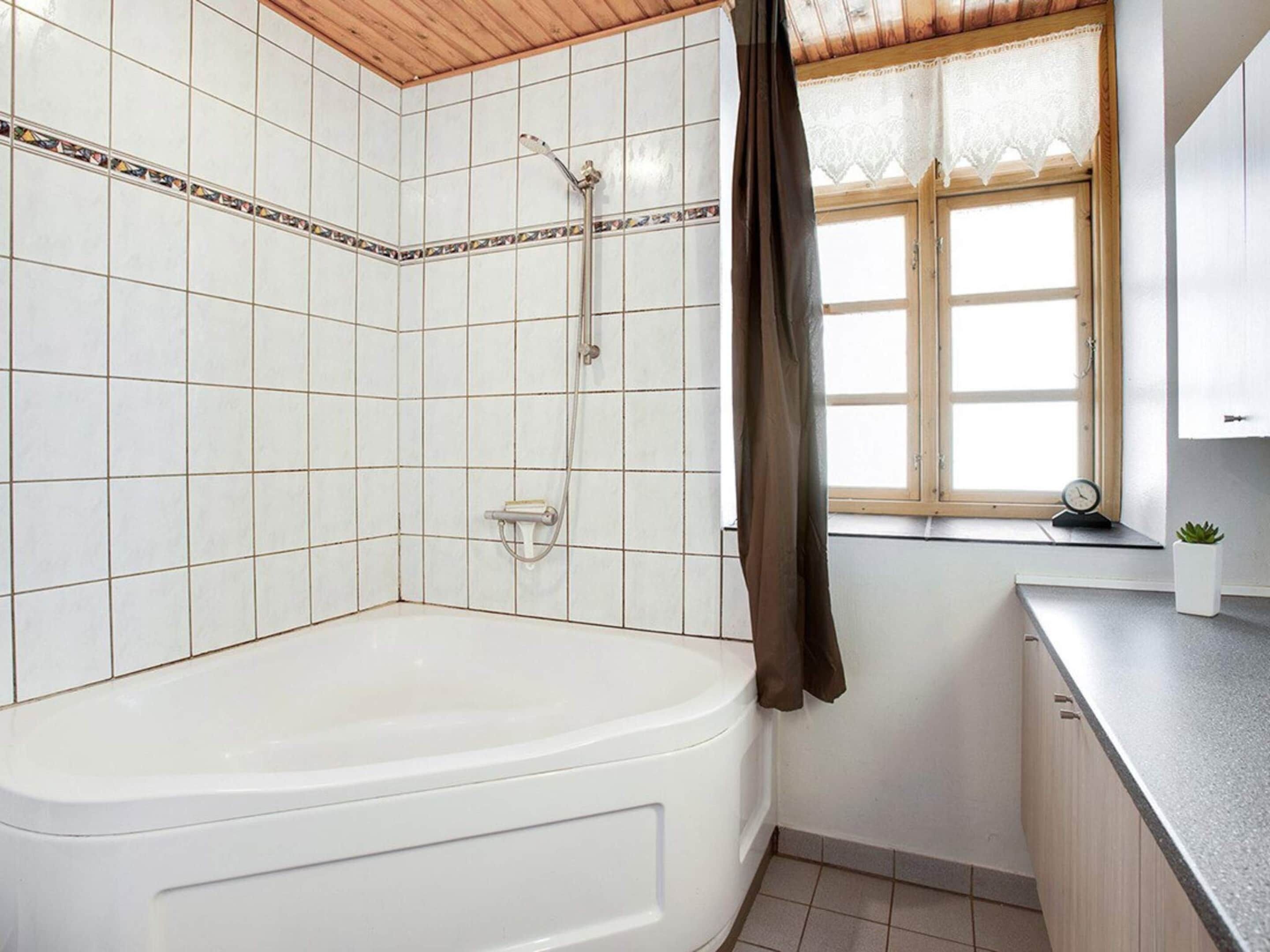 A bathroom with a bathtub, shower, tiled walls, a window with a curtain, and a small potted plant on the counter.