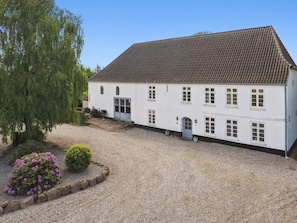 En vit byggnad i två våningar med ett tak täckt av takpannor, en grusuppfart och en liten trädgård med blommor och buskar.