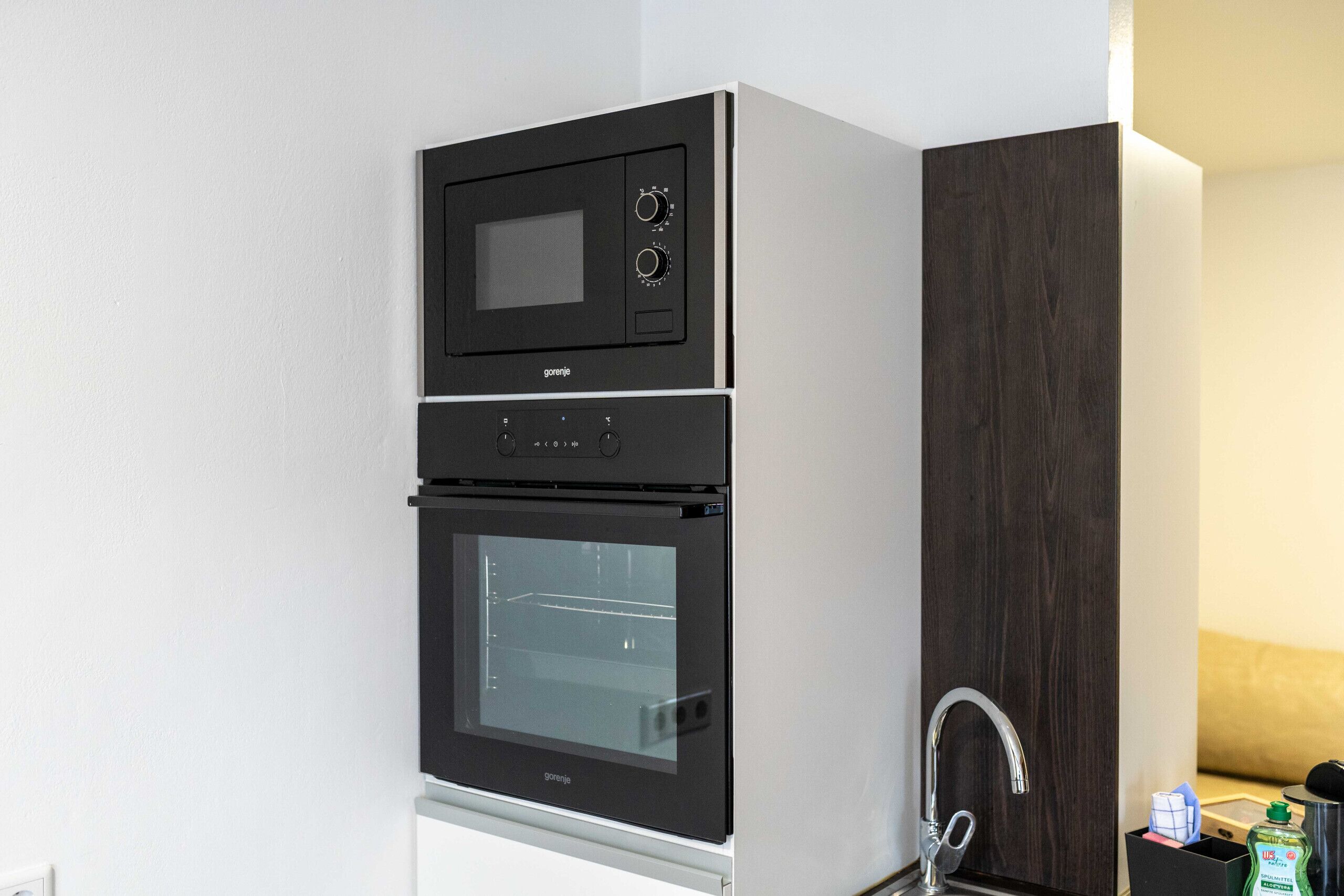 A kitchen with a built-in microwave and oven, a sink, and a wooden cabinet.