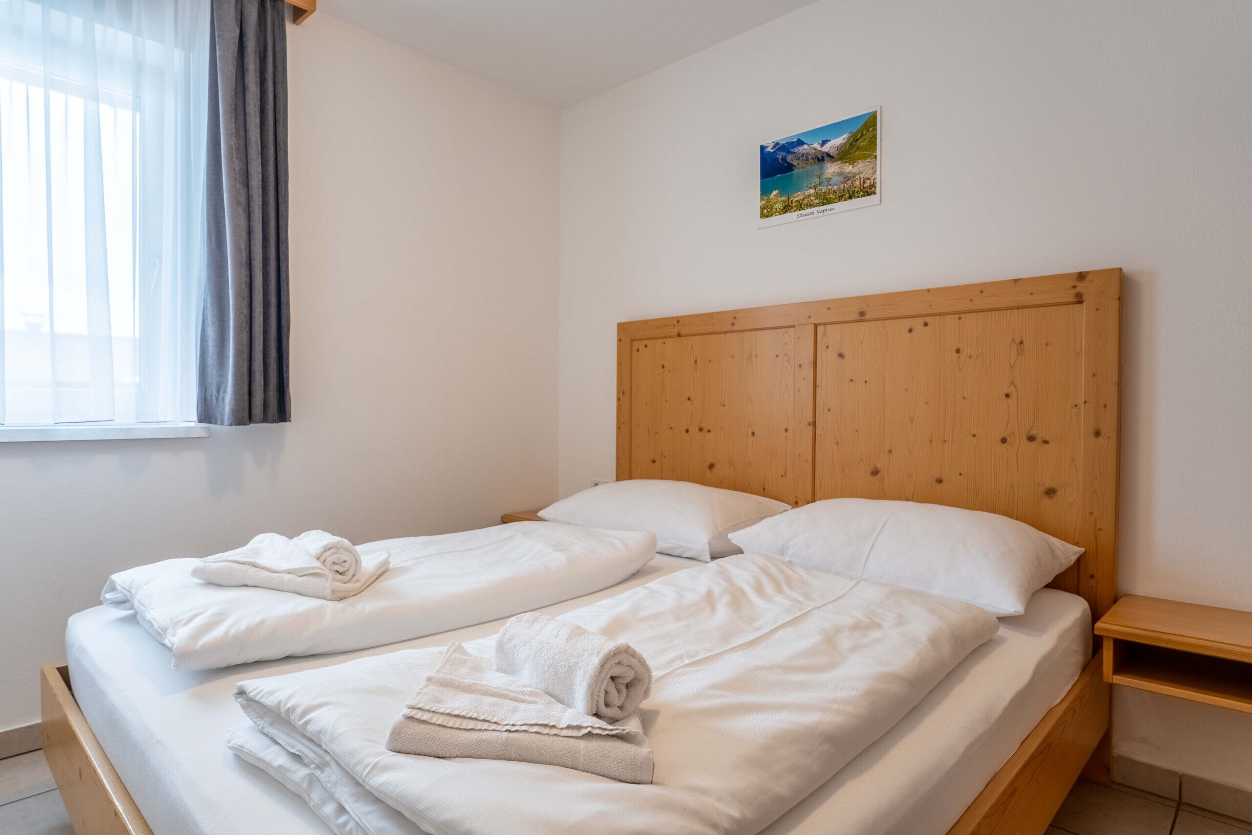 A room with a wooden headboard, two beds with white linens, a small wooden nightstand, and a window with sheer curtains.