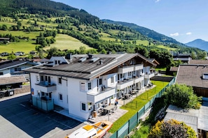 A modern hotel with multiple floors, outdoor seating, and a view of a green valley and mountains. - Maiskogelblick Apartments - TOP 12 (Piesendorf)