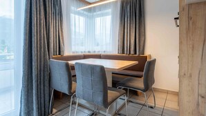 A modern dining area with a wooden table and grey upholstered chairs, large windows with sheer curtains, and a view of the outdoors. - Maiskogelblick Apartments - TOP 12 (Piesendorf)