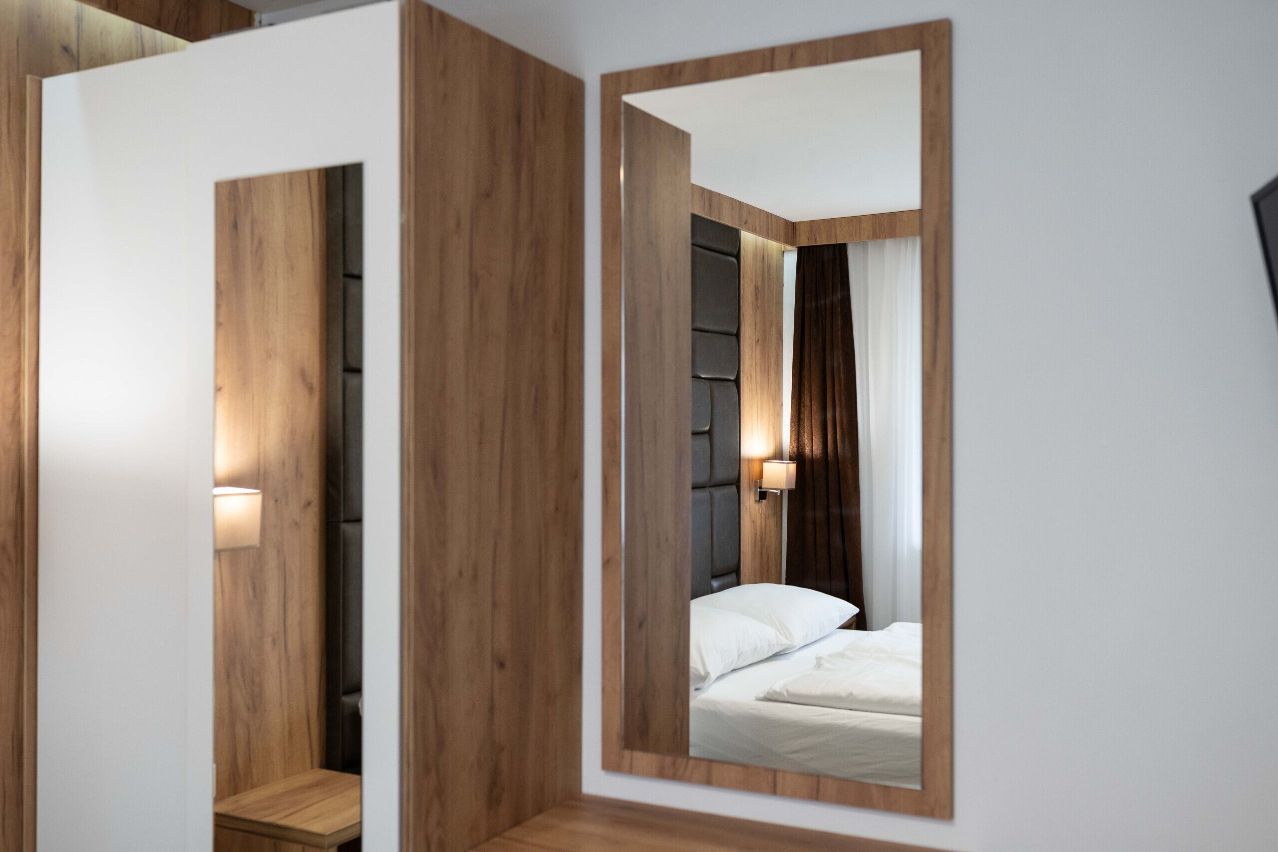 Un dormitorio moderno con un espejo grande, paneles de madera, una cama con ropa de cama blanca y una mesita de noche con lámpara.